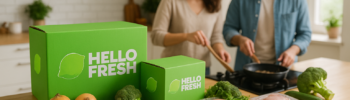 HelloFresh meal kit boxes on a kitchen counter with fresh vegetables, salmon, chicken, and recipe cards, while a couple cooks together in the background — promoting healthy eating and meal kit convenience.